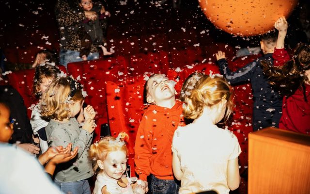 In a cinema, a group of smiling children look up at a large orange bouncy ball and foam floats through the air like snow.