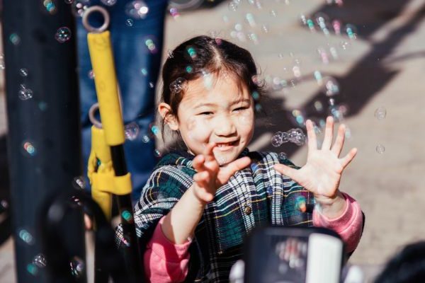 On the street on a bright day, a child smiles and reaches out her hands towards bubbles floating through the air from a bubble machine.