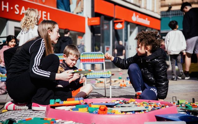 On the street on a bright day, three children surround a pink tub containing colourful pieces of LEGO, and each pick some up.