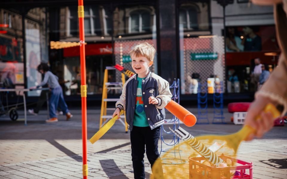 A child smiles, holding a yellow plastic racket and surrounded by colourful plastic props.