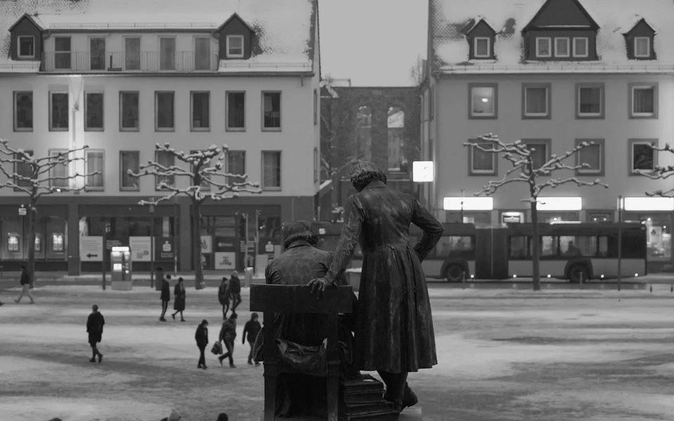 Black and white still of two statues in front of a snowy street.