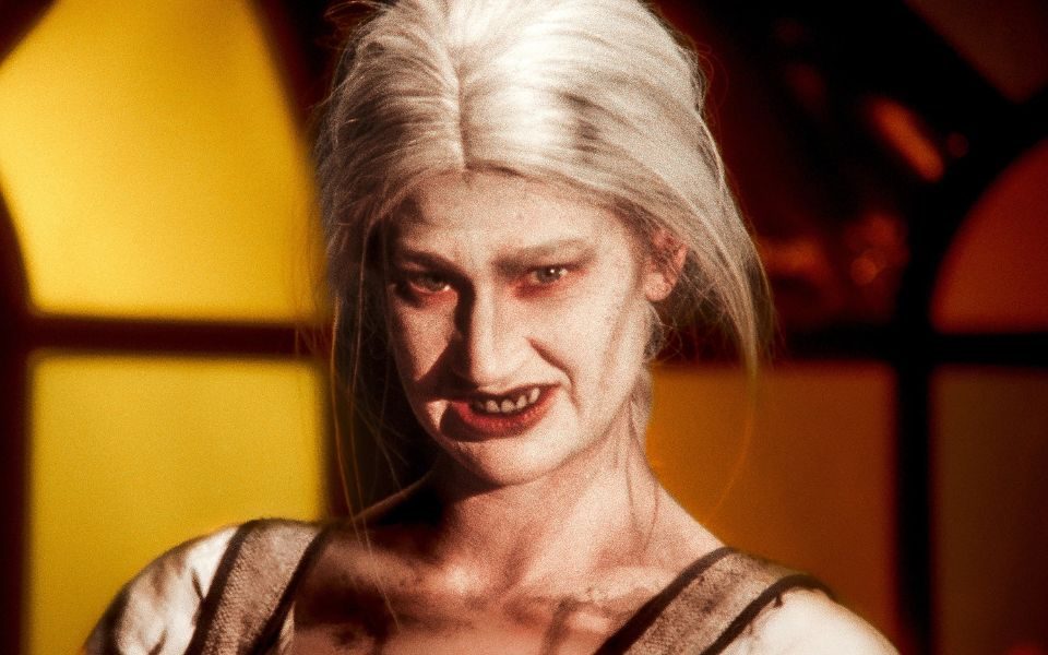 A woman with white hair and bold horror makeup grimaces, with dirt and marks visible on her torso.