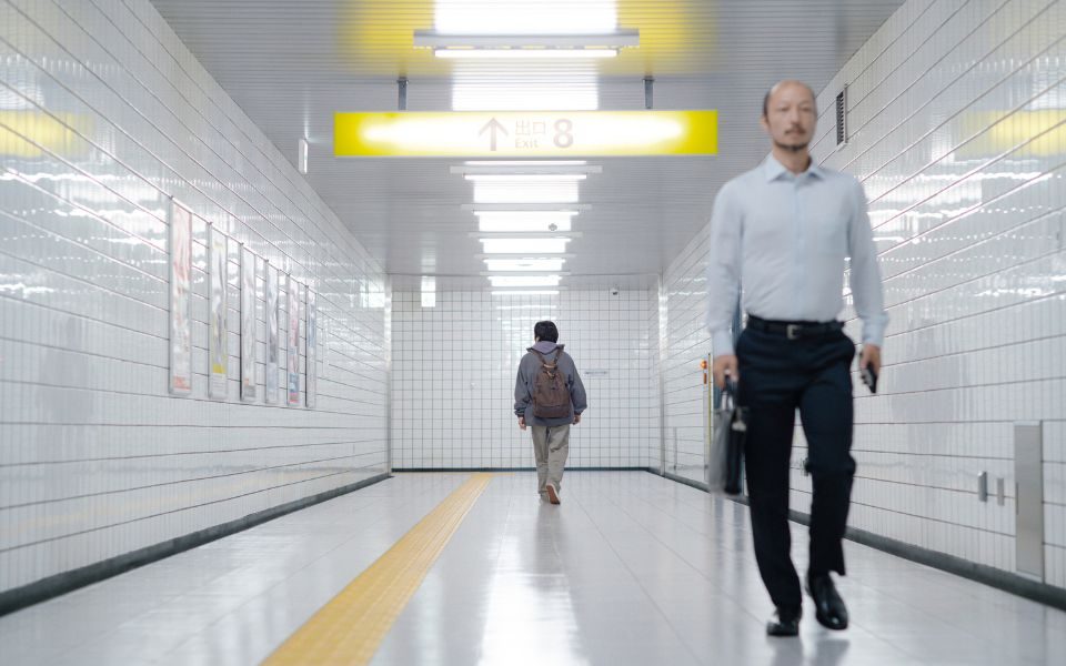 In an underground subway station corridor, with white tiles and fluorescent lights, a man in a suit walks towards the camera as someone in a coat and backpack walks in the opposition direction.