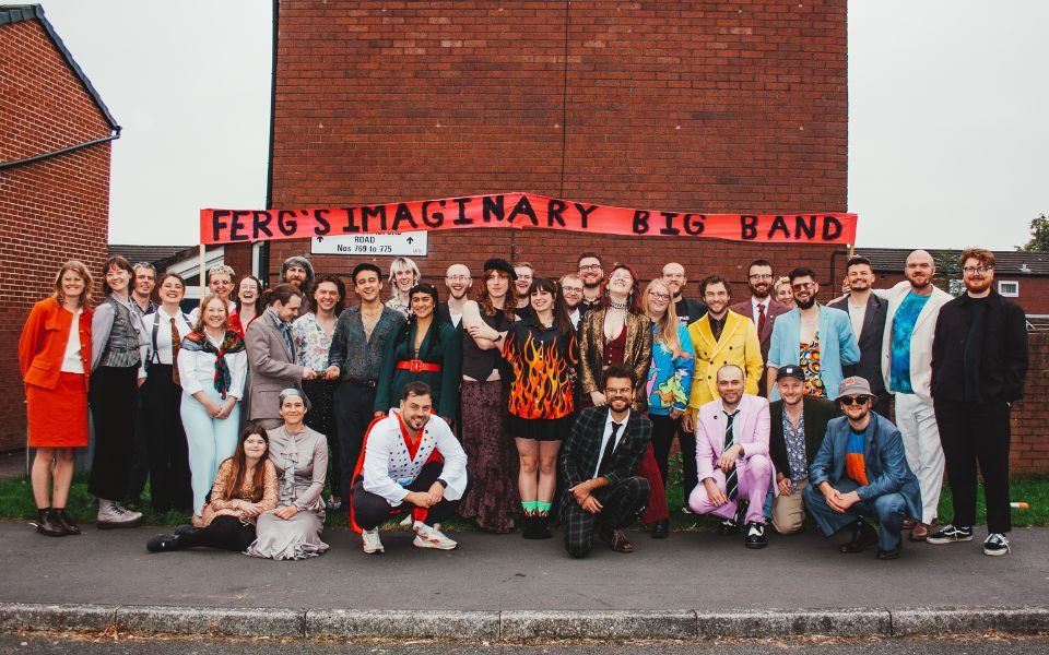 Ferg's Imaginary Big Band line up smiling on a street in front of a red brick building, adorned with a hand-painted banner that reads: Ferg's Imaginary Big Band.