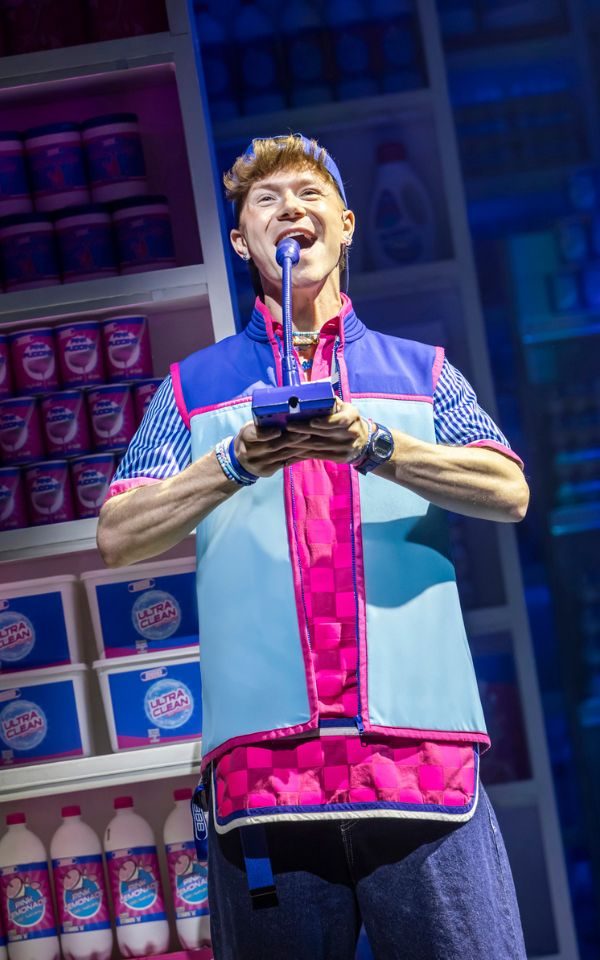 Blake Patrick Anderson speaking into a tannoy and wearing a supermarket worker uniform of pink tabard and blue fleece.