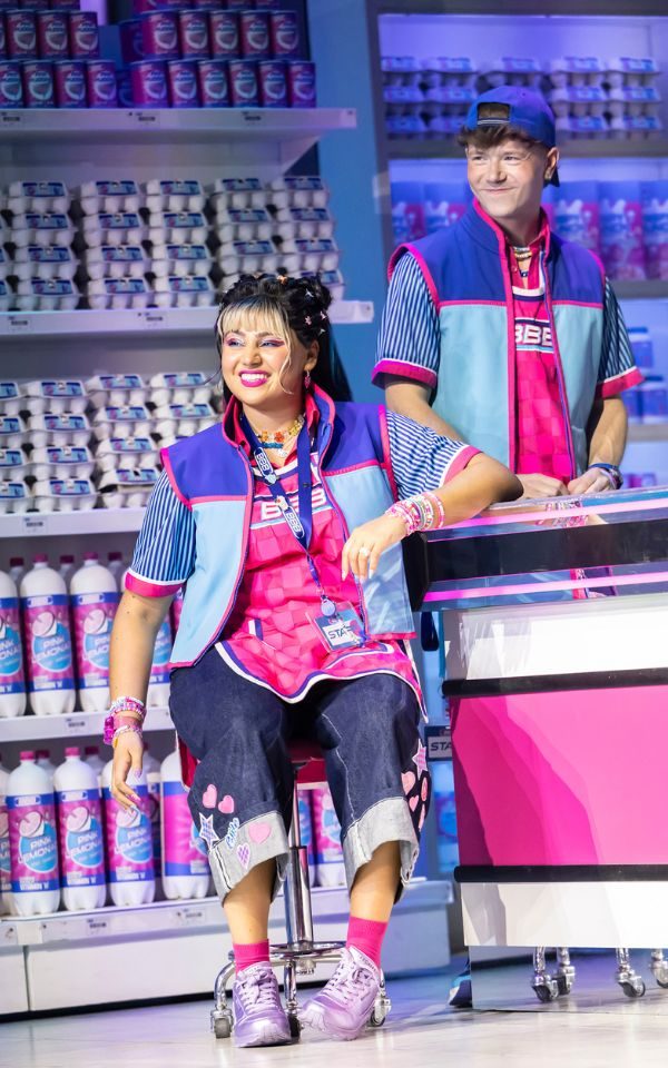 Rosie Singha as Neeta wearing a pink tabard and blue fleece as a supermarket worker uniform sits on a stool by a pink checkout counter.