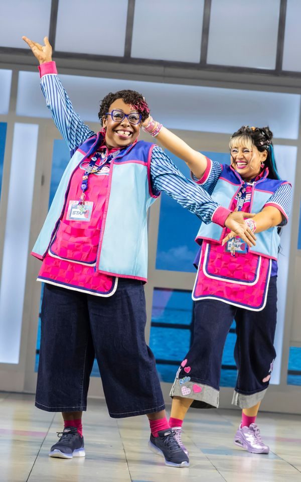 Jacqui Dubois as Vel in a supermarket worker uniform combining a pink tabard, blue fleece and lanyard. She stands smiling widely with her arms outstretched as Rosie Singha as Neeta gestures proudly towards her.
