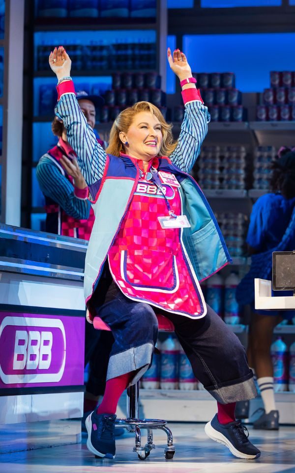 Lara Denning as Caz, wearing a bright pink tabard and blue fleece, sitting on a stool between checkout aisles and raising her arms in the air.