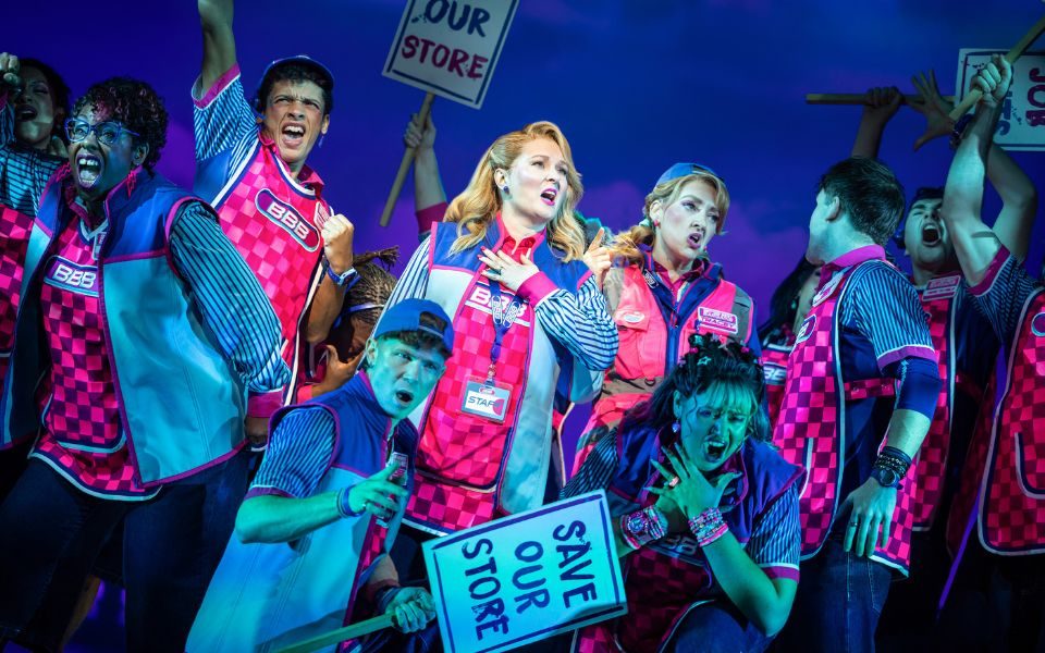 Lara Denning as Caz, wearing a pink tabard and blue fleece as a supermarket worker uniform, singing with one hand to her heart. Around her is a closeknit group of supermarket workers in the same uniform, holding protest signs reading Save Our Store.