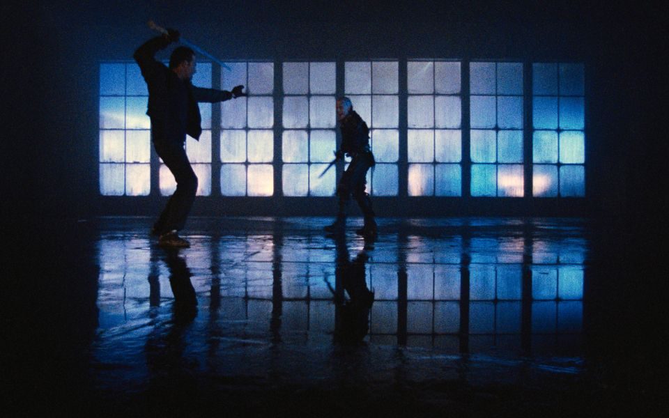 In a warehouse with a reflective, wet floor and large translucent windows, two people with swords hold fighting poses.