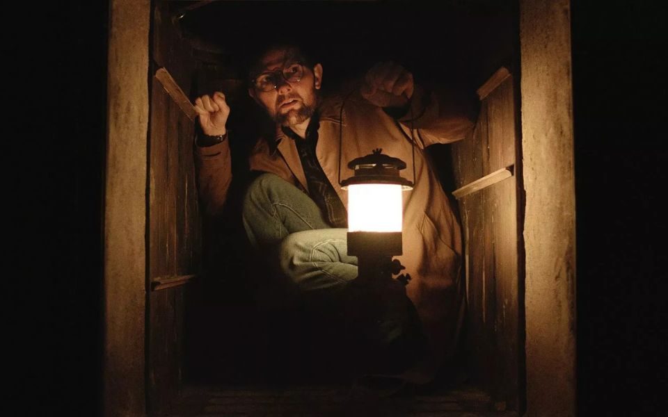A man in glasses crouches down in a tight, dark passageway made of wooden panels, holding a lantern ahead of him.