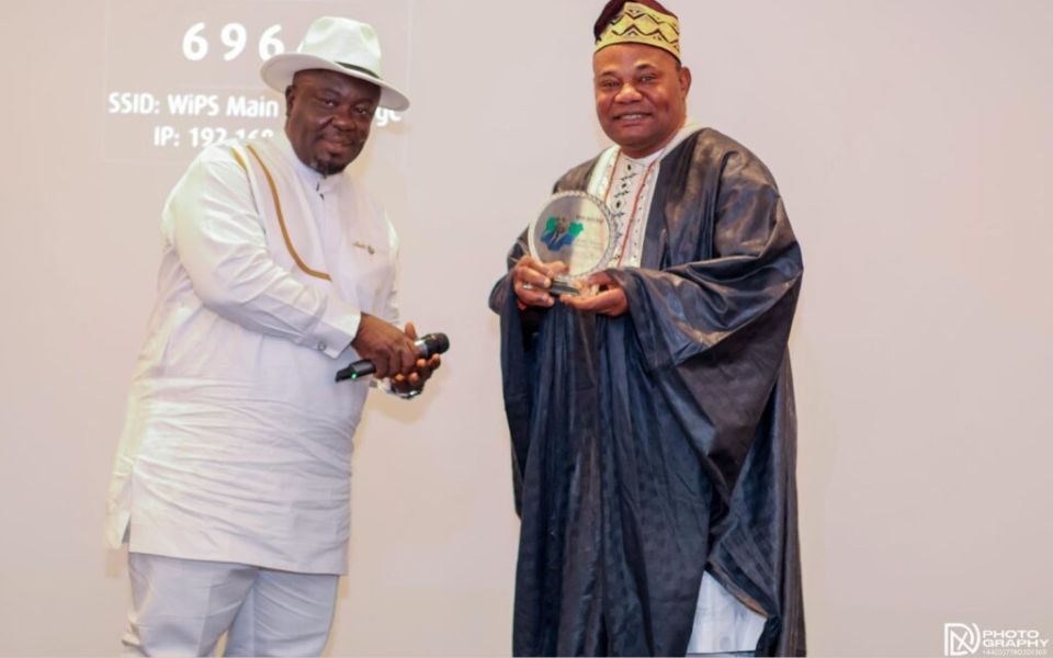 Innocent holding a transparent trophy in a gown and hat, alongside a man in a white shirt and hat, presenting the award.