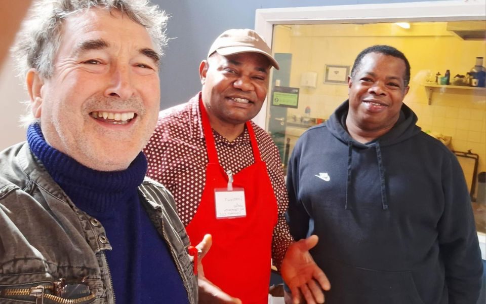 Innocent wears a cap and red apron with a name tag, alongside two other men smiling at the camera.