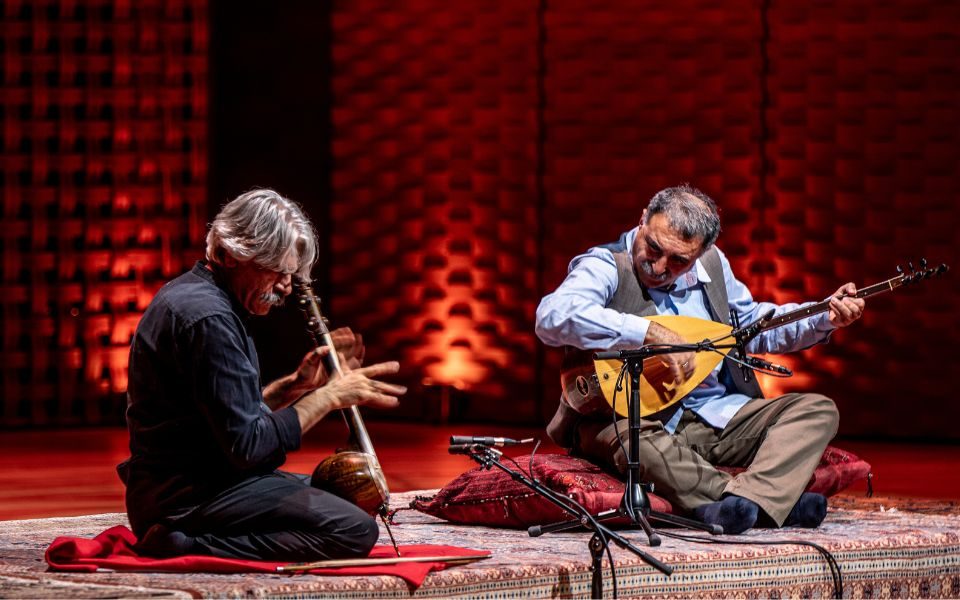 Kayhan Kalhor and Erdal Erzincan sit on cushions on a Persian rug, with their heads down as they play their instruments. They are in front of a red, textured background, lit by warm ambient lighting.