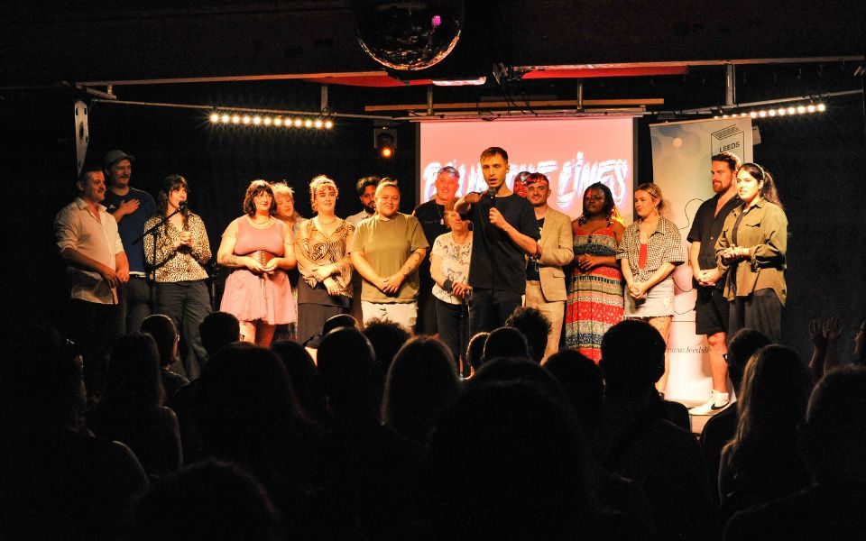 A man holds a microphone, surrounded by a large group of performers stood on stage, speaking to a crowded room.