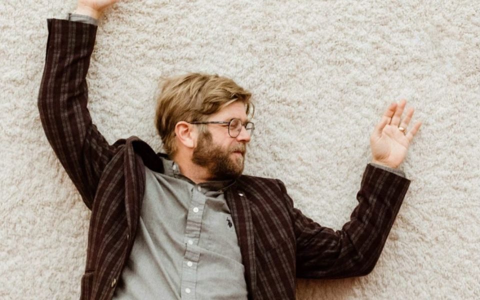 Chris Luedecke lying on a white rug, with arms outstretched and eyes closed. He wears glasses, a grey button-up shirt and chequered blazer.