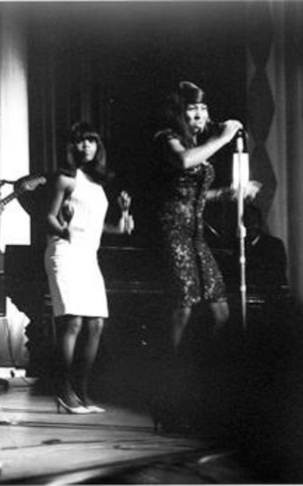 Black and white photo of PP Arnold dancing in a white dress and shaking maracas, behind Tina Turner in a sequined dress, singing into a microphone on-stage.