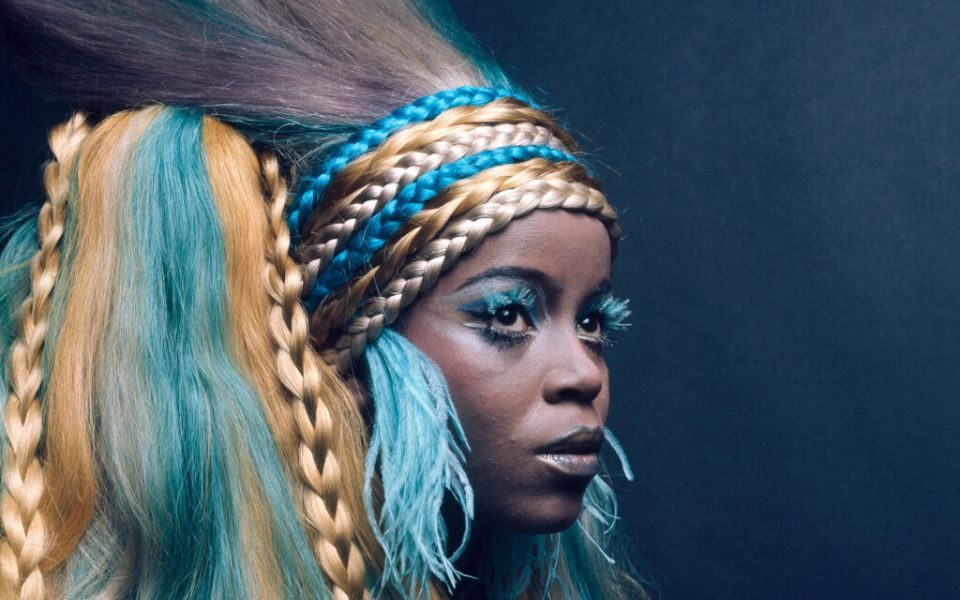 PP Arnold wearing an elaborate headpiece of golden and blue plaited hair, long feathered earrings and blue eyeshadow.