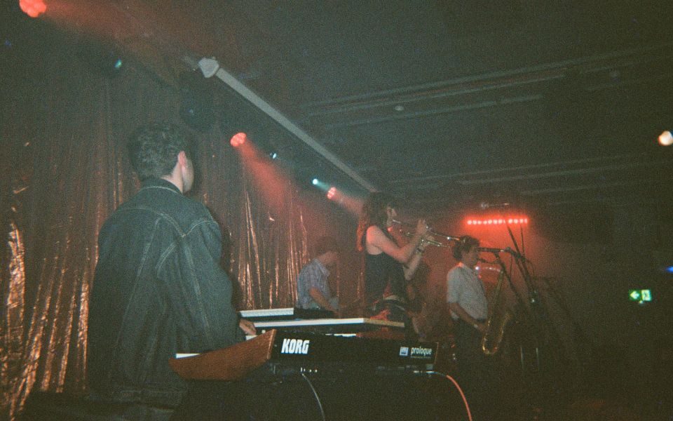 On stage in front of a sparkly curtain, a band performs. There is a keyboard player, two guitarists, a trombone player and saxophonist.