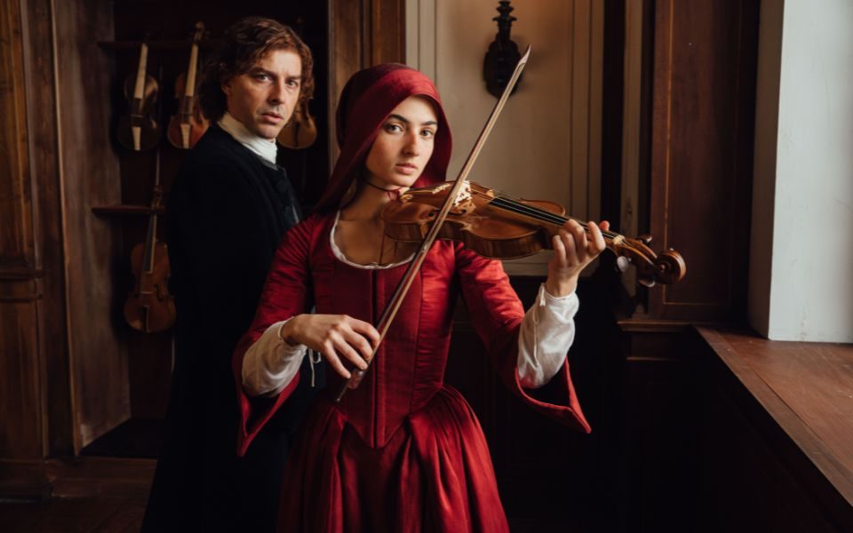 A young woman in a red dress and period headwear plays a violin, beside a stern man in traditional black clothing. Other instruments are visible in a wooden wardrobe in the background.