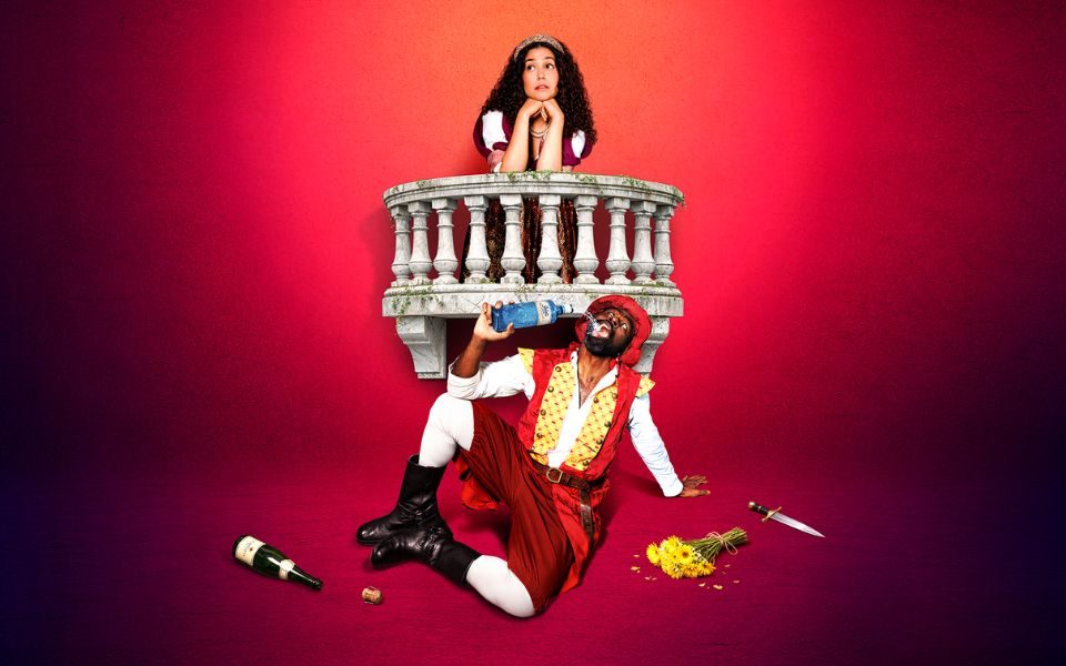 Stood at a prop balcony, an actress in period costume rests her head in her hands and looks off-camera. In front of her, another actor is sat on the floor in a red tunic and boots, drinking from a bottle of gin beside an empty bottle and discarded dagger.