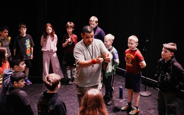 Testament stands in the centre of a group of young people on-stage, gesturing two thumbs up to the group.