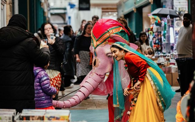 A dancer in an elaborate dress crouches down with a colourful puppet elephant in a busy market, capturing the attention of a child.