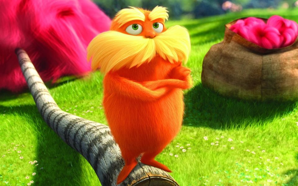 Vibrant animated still. The Lorax, with orange fur and a large yellow moustache, crosses his arms and stands on a log.