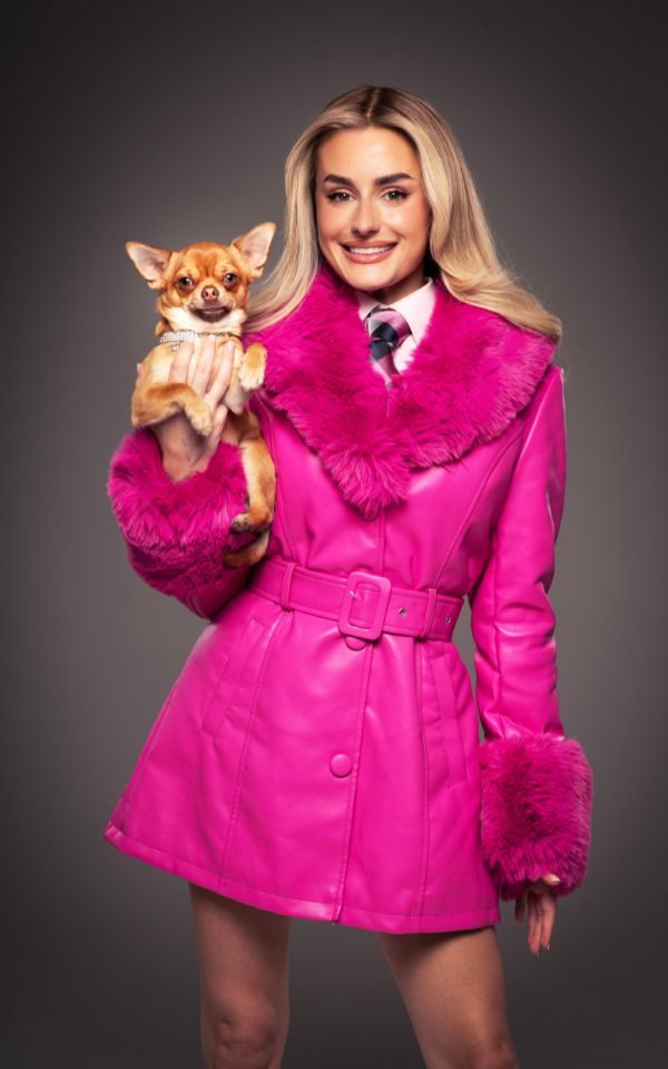 Amber Davies with blonde hair in a hot pink belted leather coat with a pink fur collar. She smiles at the camera and holds up a light brown chihuahua.