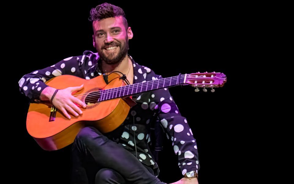 A man smiles on a stool onstage, wearing a polka dot shirt and holding an acoustic guitar.