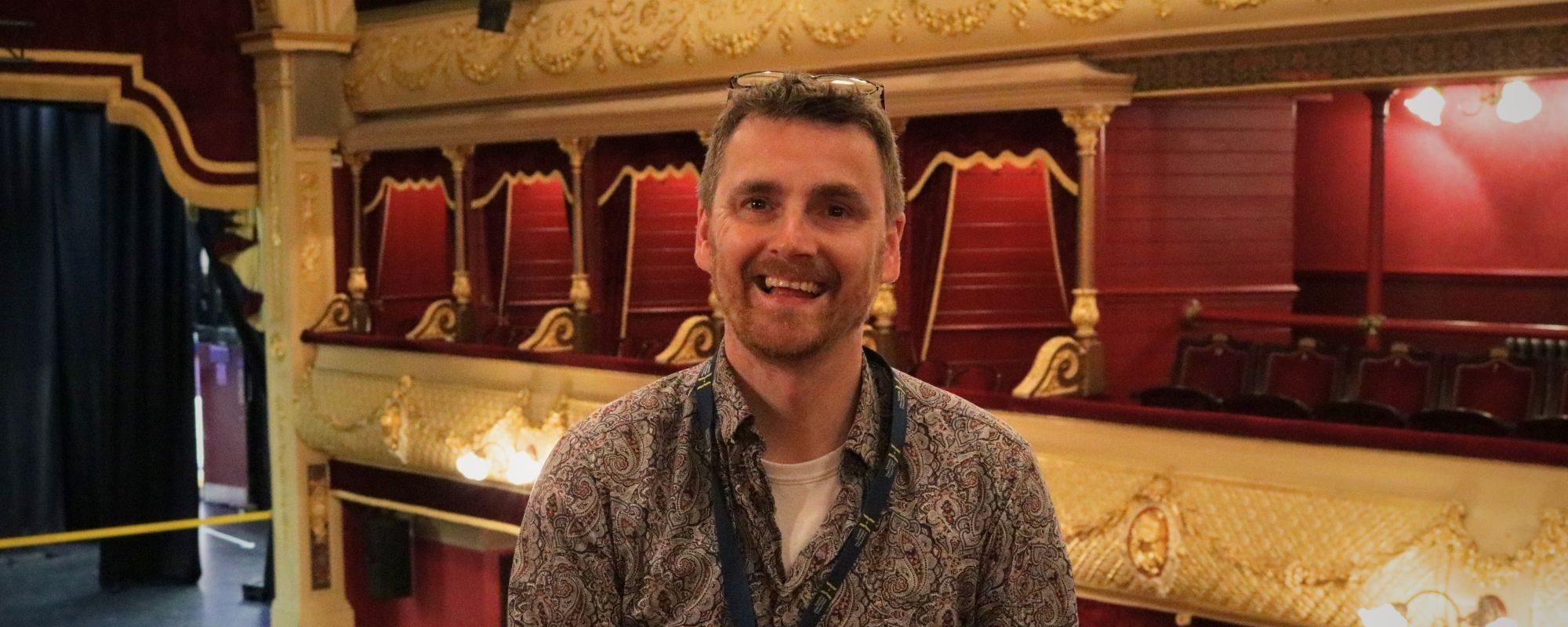 Frank smiles, stood in the Circle at City Varieties Music Hall, the auditorium lit behind him