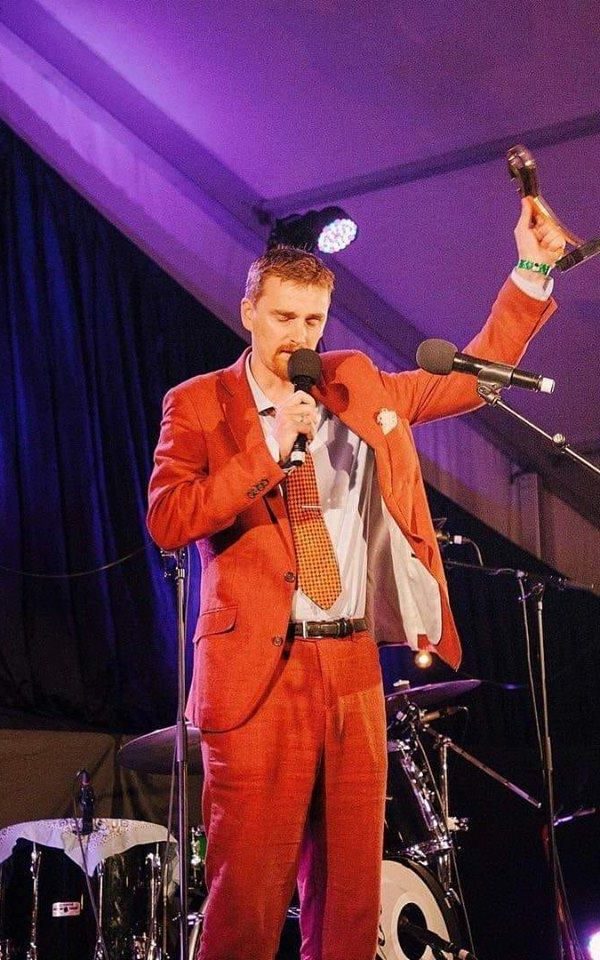 Frank on stage in an orange suit, speaking into a microphone and holding up an award