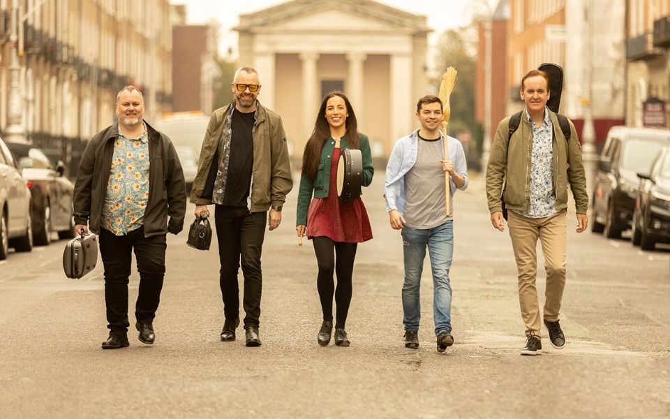 On a sunny street, the members of Irish House Party smile and walk towards the camera, each carrying an instrument case, one holding a broom.