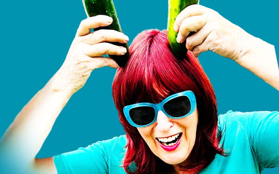 Janet Street Porter with bright red hair and blue sunglasses, laughing and holding up two cucumbers like horns above her head.