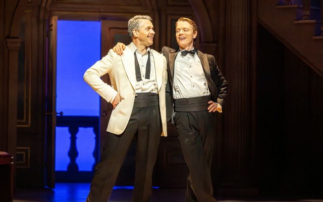 On stage, two men in tuxedo smile at each other and stand proudly, with one putting his arm around the other's shoulder.