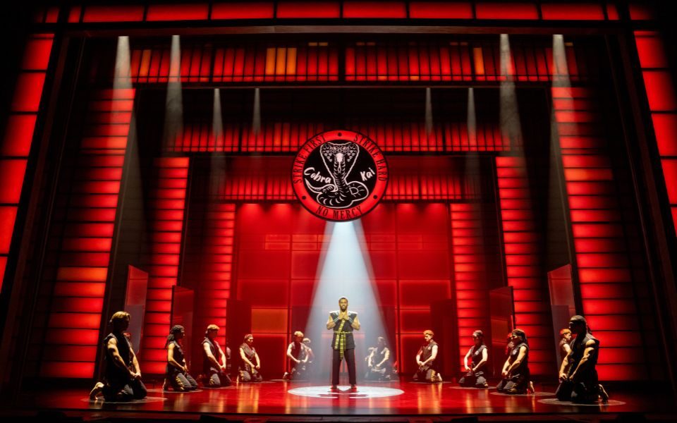 On a dark red stage, a cast sit in karate uniforms in a circle, a spotlight on their teacher stood centre stage