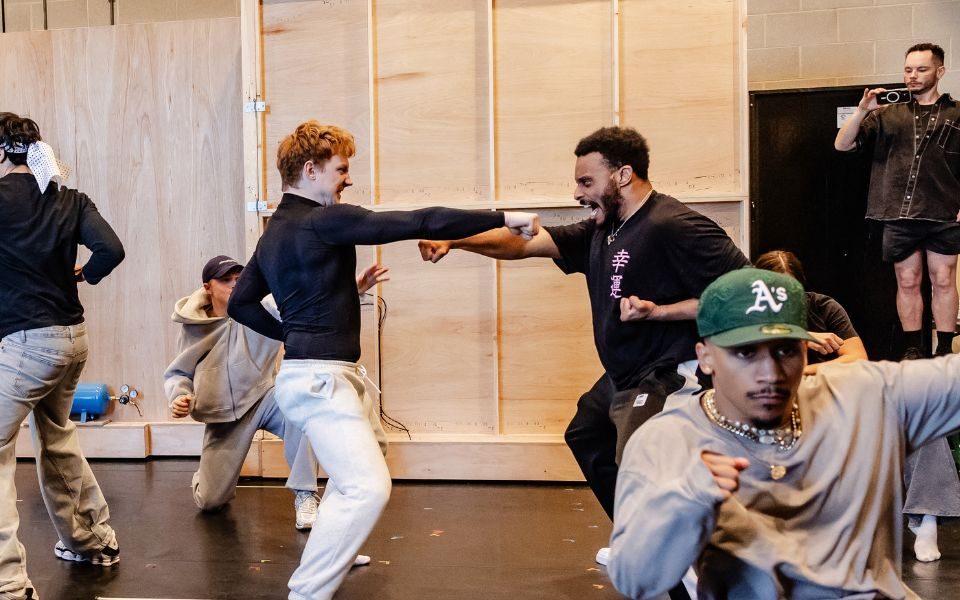 Two cast members dramatically punch towards one another, in a rehearsal room