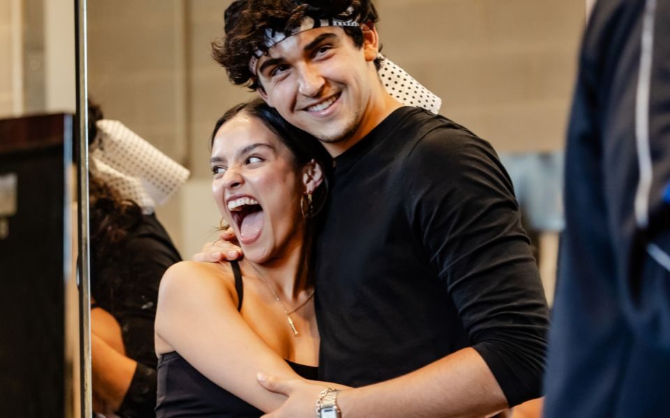 Two cast members smile as they hug each-other in a rehearsal room