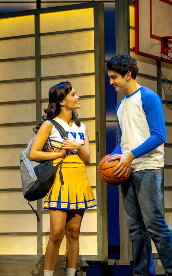 Two cast members smile at each-other on stage, holding a backpack and a basketball