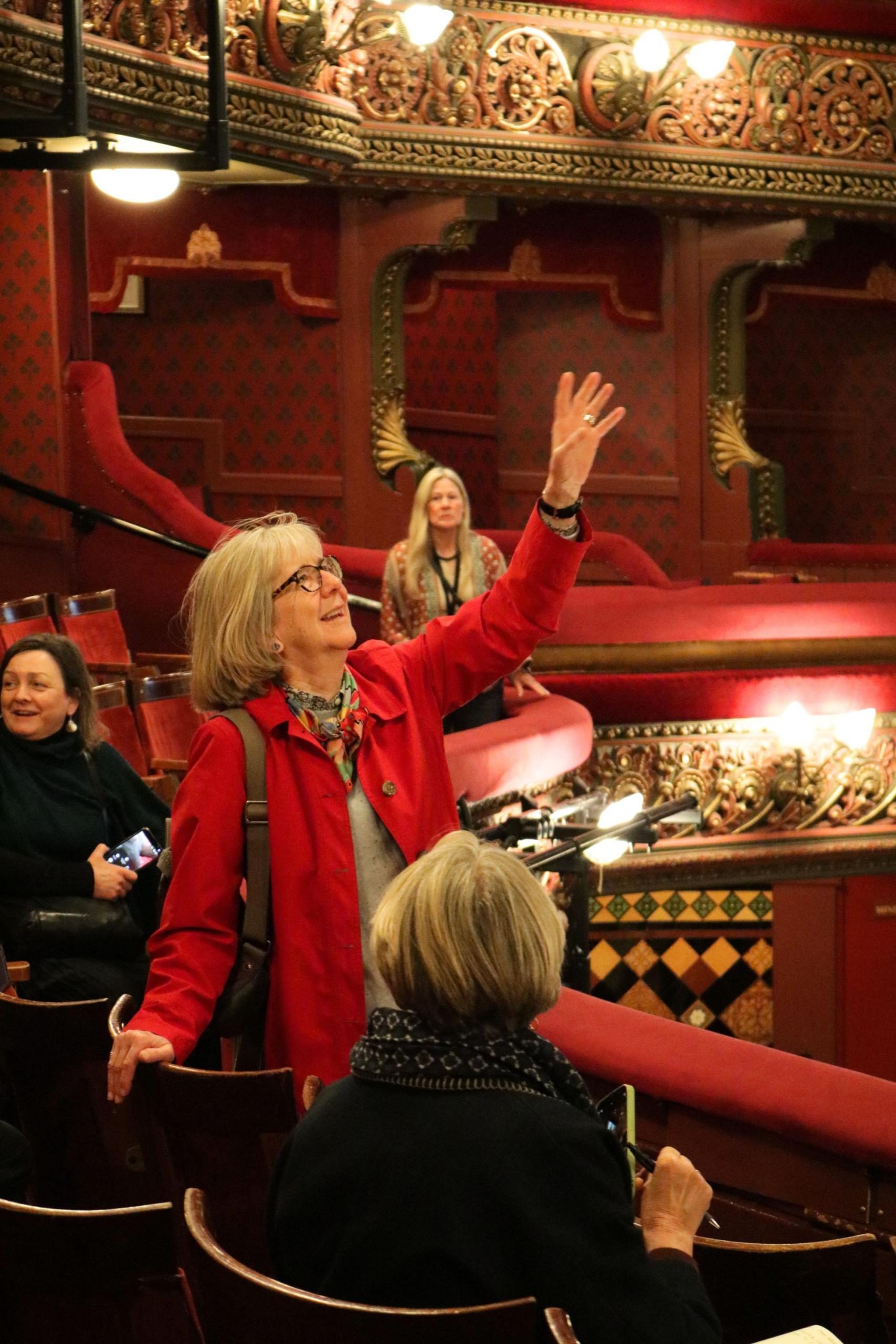 Stood next to seats in the dress circle. a person in a red coat smiles, reaches out a hand and looks up across the auditorium of the Leeds Grand Theatre auditorium.
