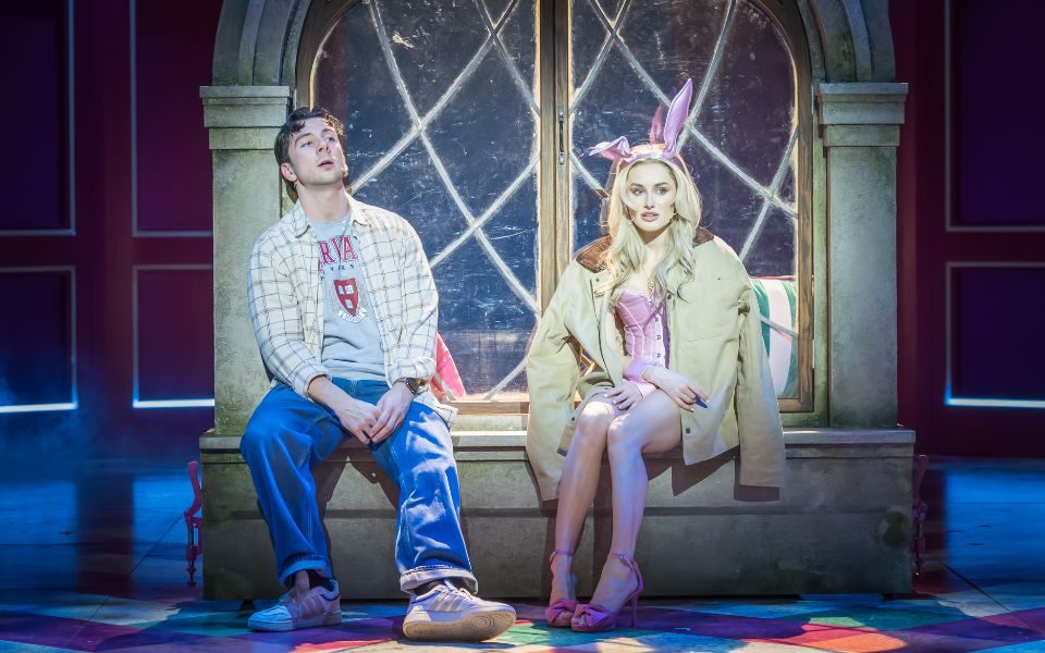 A man in jeans and a check shirt and a woman in a baby pink Playboy Bunny costume and oversized beige coat sit next to each other by a large window.