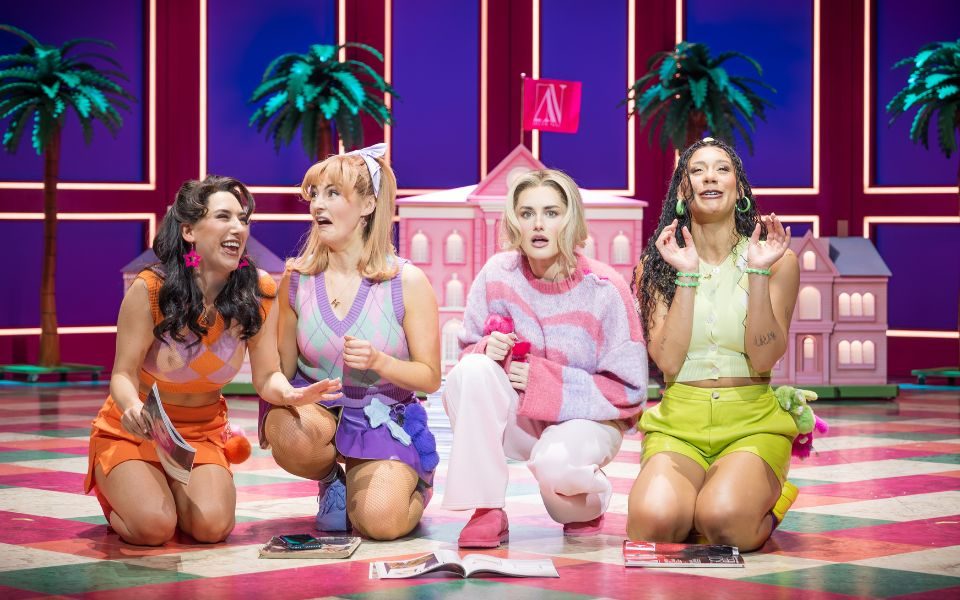 Four women kneeling in a line. They each wear preppy sorority outfits in orange, purple, pink and green respectively. In front of them are a collection of magazines and behind them is a model version of a pink university building.