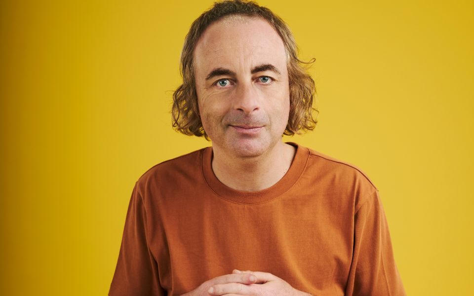 Paul Foot smirks and clasps his palms together, wearing an orange t-shirt in front of a yellow backdrop.