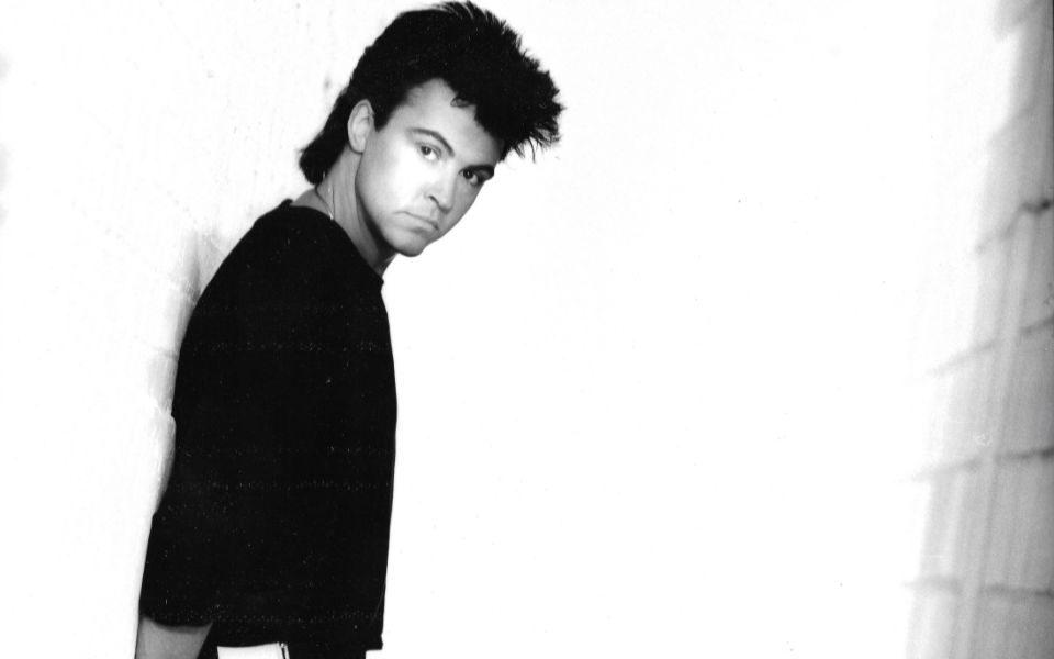 Black and white photo of Paul Young leaning against a wall, wearing a long sleeve black t-shirt.