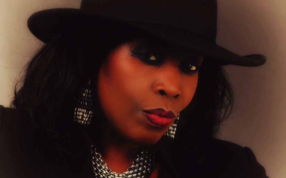 Ruby Turner posing in front of a plain backdrop, wearing silver jewellery, a black outfit and brimmed hat.