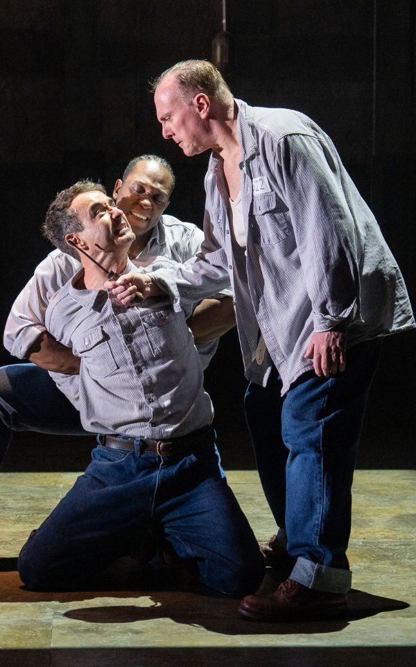 Joe McFadden in The Shawshank Redemption kneels down while a man holds his hands behind his back and another man holds a knife to his throat.