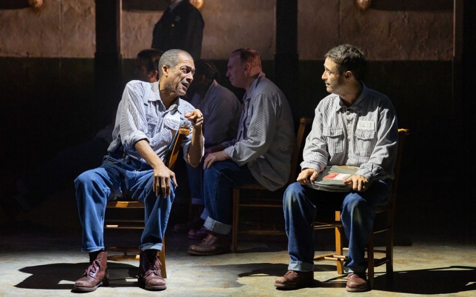 Ben Onwukwe and Joe McFadden sitting on wooden chairs and talking to each other. They wear matching grey shirts and blue jeans as prison outfits.