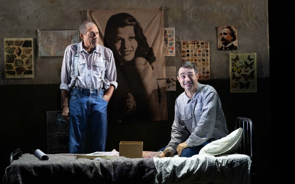 Joe McFadden sitting on a prison bed next to a small box. Behind him, Ben Onwukwe stands watching him next to a large poster of Rita Hayworth.