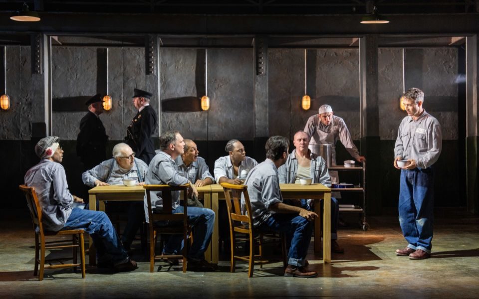 Joe McFadden as Andy Dufresne holding a bowl and standing in front of a table where a number of prison inmates sit staring at him in a prison kitchen.