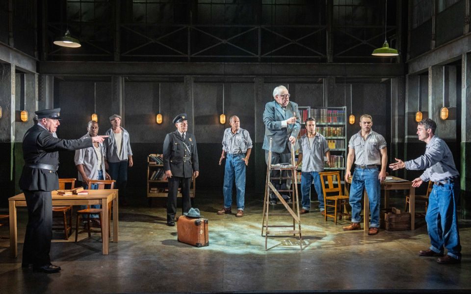 A prison warden points across a prison reading room to Joe McFadden as Andy Dufresne who looks at him pleadingly. A elderly man stands at the top of a ladder while other prison inmates around the room watch the situation.